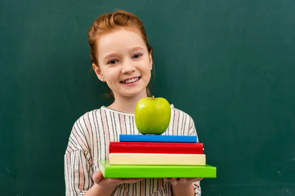 Shoolgirl jengibre alegre sosteniendo libros y manzana verde en frente de pizarra en el aula - foto de stock