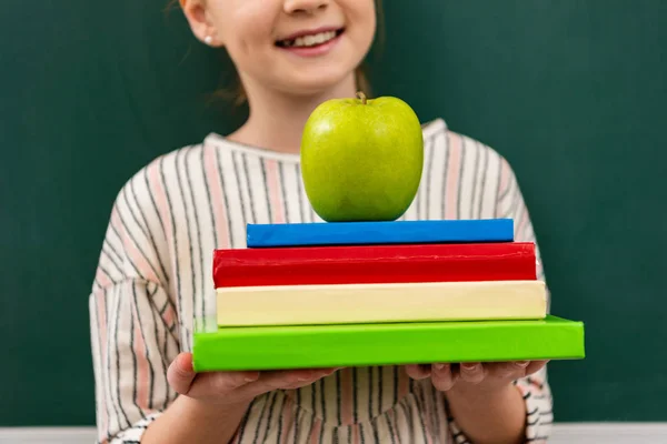 Cropped view of cheerful ginger shoolgirl holding books and green apple in front of blackboard in classroom — Stock Photo