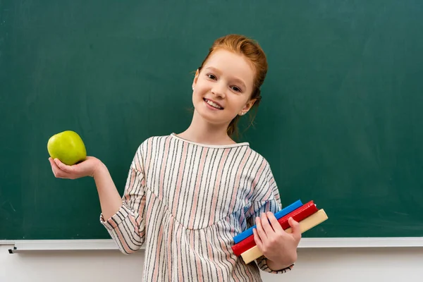 Cheerful ginger shoolgirl holding books and green apple in front of blackboard in classroom — Stock Photo
