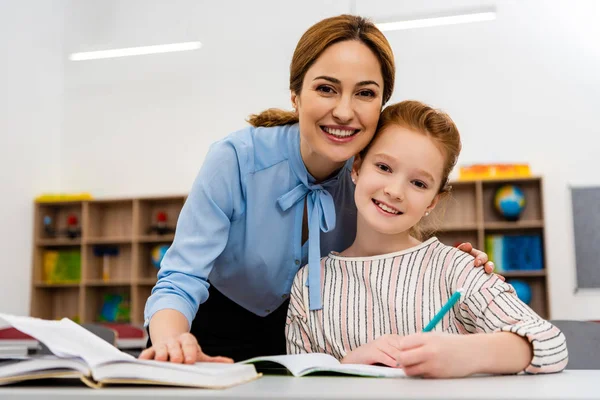 Lächelnder Lehrer in blauer Bluse umarmt Schüler und blickt in die Kamera — Stockfoto
