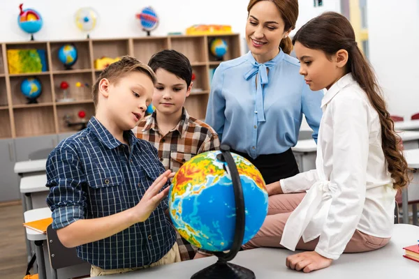 Profesor y alumnos tocando globo con sonrisa mientras estudian geografía en el aula - foto de stock
