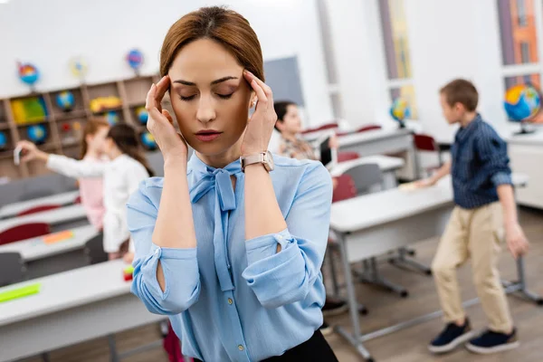 Profesora cansada en blusa azul de pie frente a escritorios y rostro conmovedor - foto de stock