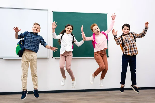 Four pupils with backpacks jumping in front of blackboard — Stock Photo