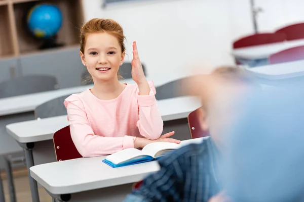 Smiling schoolgirl with book raising hand up during lesson in classroom — Stock Photo