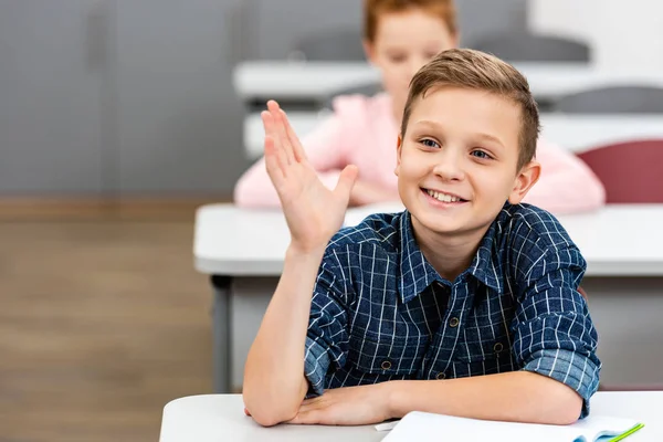 Écolier en chemise à carreaux levant la main pendant la leçon en classe — Photo de stock