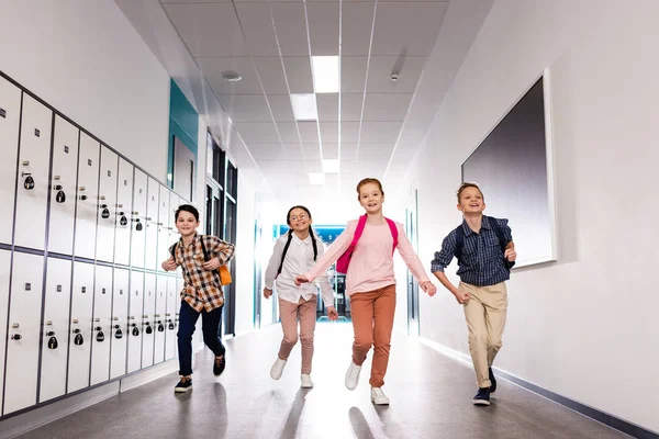 Cuatro alumnos emocionados con mochilas corriendo corredor después de clases - foto de stock