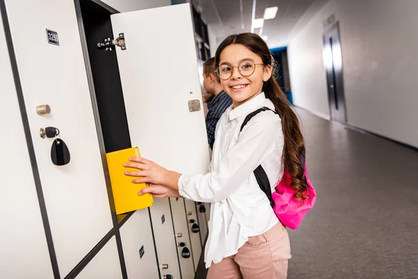Fröhliche Schülerin mit Brille legt Buch während der Schulpause ins Schließfach — Stockfoto