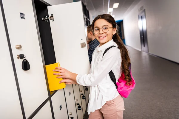 Joyful schoolgirl in glasses putting book in locker during brake in school — Stock Photo