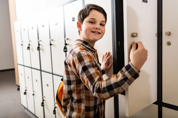 Улыбающийся школьник в клетчатой рубашке с рюкзаком, открывающим шкафчик — стоковое фото