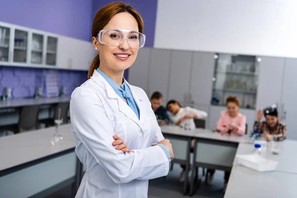 Profesora sonriente con gafas protectoras de pie con los brazos cruzados delante de los alumnos durante la lección de química - foto de stock