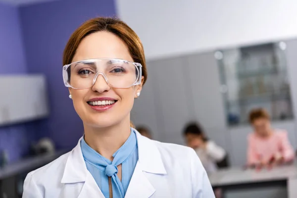 Professor sorridente em óculos de proteção em pé na frente dos alunos durante a aula de química — Fotografia de Stock