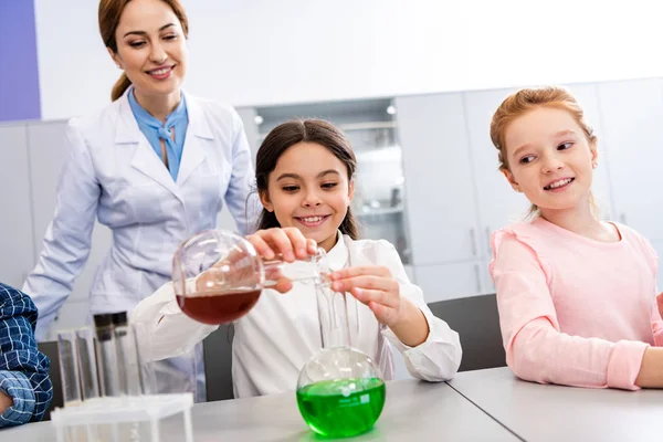 Colegiala sonriente con vasos haciendo experimento químico durante la lección de química - foto de stock