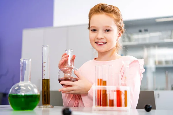 Colegiala sonriente sosteniendo el vaso de precipitados y mirando a la cámara durante la lección de química - foto de stock