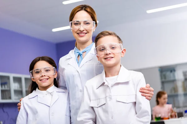 Smiling teacher in white coat and protective goggles embracing pupils during chemistry lesson — Stock Photo