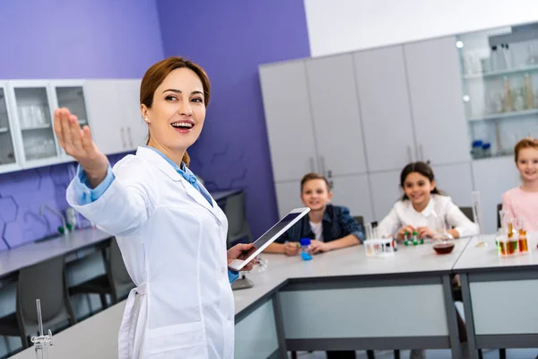 Profesor de química sonriente en bata blanca sosteniendo tableta digital y señalando con la mano - foto de stock