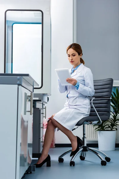 Professora de química concentrada em casaco branco usando tablet digital enquanto sentada em poltrona em sala de aula — Fotografia de Stock