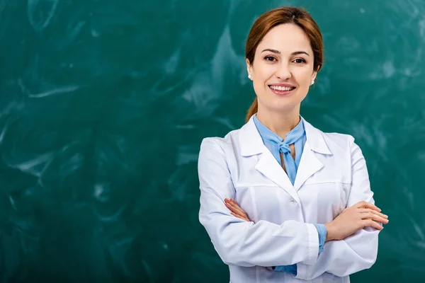 Smiling chemistry teacher in white coat standing with crossed arms in front of blackboard — Stock Photo
