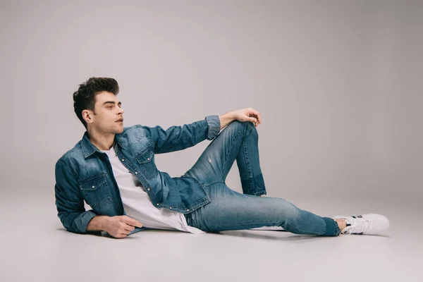 Handsome man in jeans, skirt and t-shirt lying on floor and looking away on grey background — Stock Photo