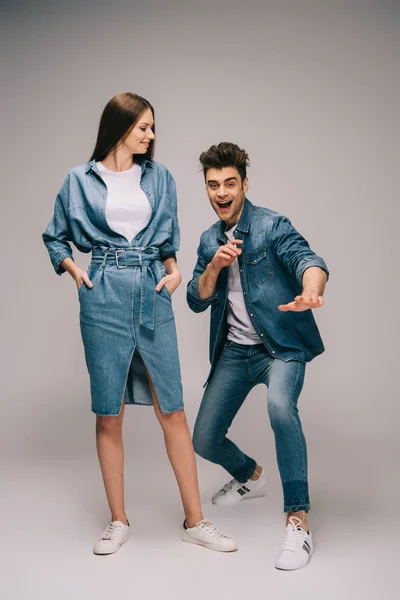 Дівчина в джинсовій сукні з руками в кишенях і усміхнений хлопець в джинсах і сорочці дивиться на камеру — стокове фото