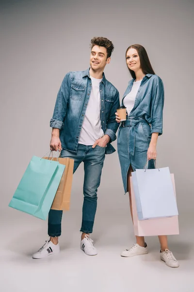 Подружка в джинсовом платье с бумажной чашкой и красивым парнем в джинсах и рубашке с сумками для покупок — стоковое фото