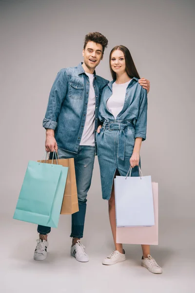 Novia en vestido de mezclilla y novio guapo en jeans y camisa sosteniendo bolsas de compras y mirando a la cámara - foto de stock