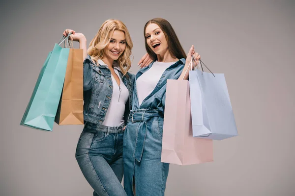 Hermosa rubia y feliz morena mujeres en ropa de mezclilla sosteniendo bolsas de compras y sonriendo - foto de stock