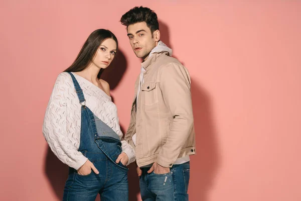 Brunette and beautiful girlfriend in denim overalls and handsome boyfriend with hands in pockets looking at camera — Stock Photo