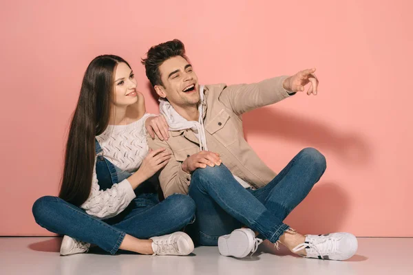 Brunette and beautiful girlfriend in denim overalls and handsome boyfriend sitting on floor and pointing with finger — Stock Photo