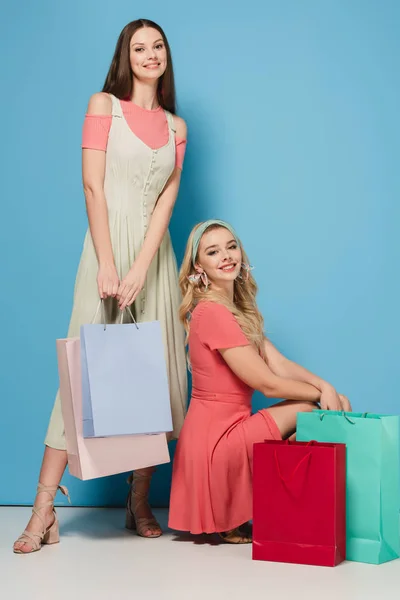 Morena sonriente y mujeres rubias en vestidos sosteniendo bolsas de compras y mirando a la cámara - foto de stock