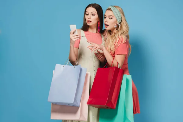 Здивована брюнетка і блондинка в сукнях тримає сумки і використовує смартфон — стокове фото