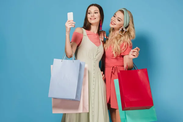 Morena sonriente y mujeres rubias en vestidos sosteniendo bolsas de compras y tomando selfie - foto de stock