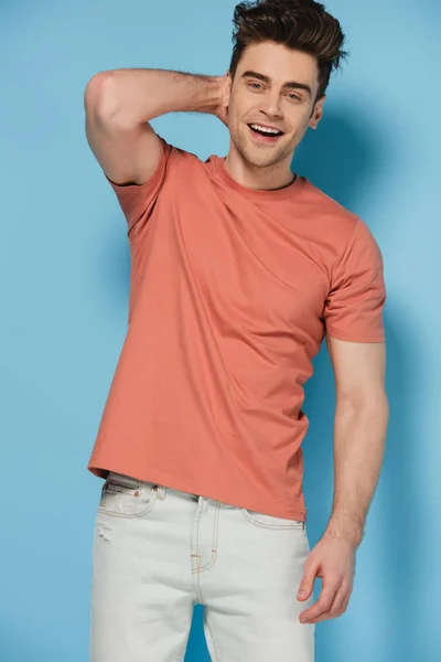 Schöner und brünetter Mann im T-Shirt, der in die Kamera schaut und auf blauem Hintergrund lächelt — Stockfoto