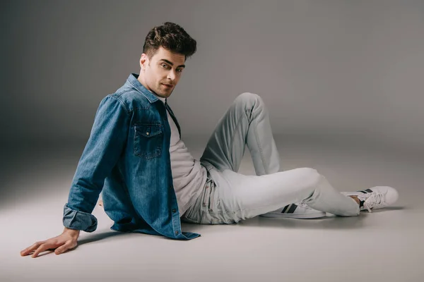 Hombre guapo en camisa de mezclilla y jeans sentado en el suelo y mirando a la cámara — Stock Photo