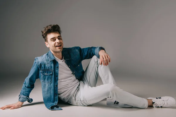 Lächelnder gutaussehender Mann in Jeanshemd und Jeans sitzt auf dem Boden und blickt in die Kamera — Stockfoto