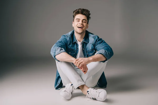 Beau homme souriant en denim chemise et jeans assis sur le sol et regardant la caméra — Photo de stock