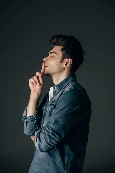 Vista lateral do homem bonito e morena em camisa jeans mostrando shh gesto isolado em preto — Fotografia de Stock