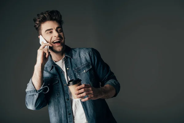 Sonriente y morena hombre en camisa de mezclilla sosteniendo taza de papel y hablando en teléfono inteligente aislado en negro - foto de stock