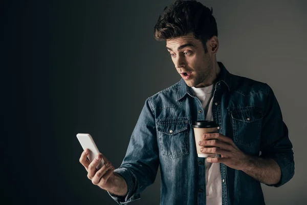 Homme beau et choqué en denim chemise tenant tasse en papier et smartphone sur fond noir — Photo de stock