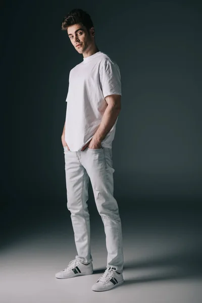 Hombre guapo en camiseta blanca y jeans con las manos en los bolsillos mirando a la cámara - foto de stock