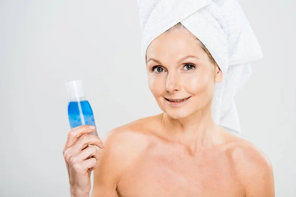 Attractive and smiling mature woman in towel holding bottle with micellar water and looking at camera — Stock Photo