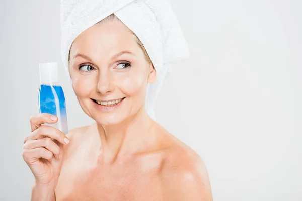 Atractiva y sonriente mujer madura en botella de toalla con agua micelar y mirando hacia otro lado - foto de stock