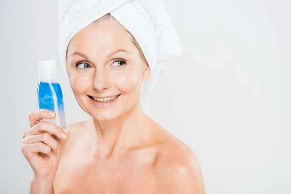 Attraktive und lächelnde reife Frau im Handtuch, Flasche mit Wasser im Keller haltend und wegschauend — Stockfoto