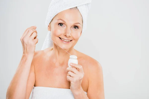 Привлекательная и улыбающаяся зрелая женщина в полотенце держит зубную нить и смотрит в камеру — стоковое фото