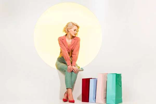 Bela menina elegante sentado no círculo amarelo perto de sacos de compras e posando no branco — Fotografia de Stock