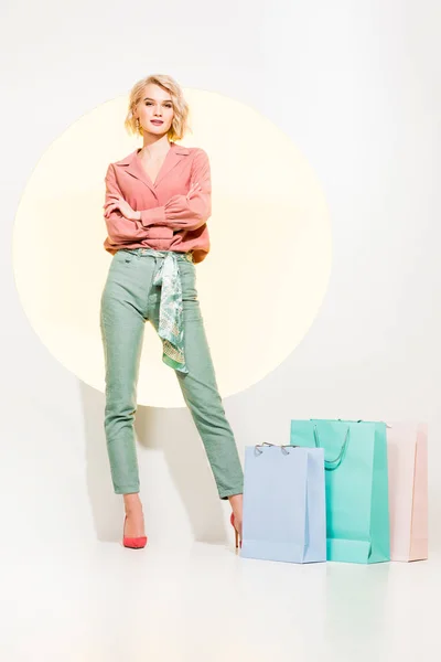 Hermosa chica con estilo con los brazos cruzados posando cerca de bolsas de compras en blanco con círculo amarillo - foto de stock