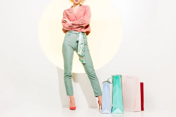 Vista cortada de menina elegante com braços cruzados posando perto de sacos de compras no branco com círculo amarelo — Fotografia de Stock
