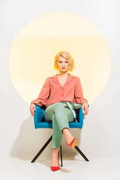 Belle fille élégante dans des vêtements colorés regardant l’appareil-photo et s’asseyant dans le fauteuil sur le blanc avec le cercle jaune — Photo de stock