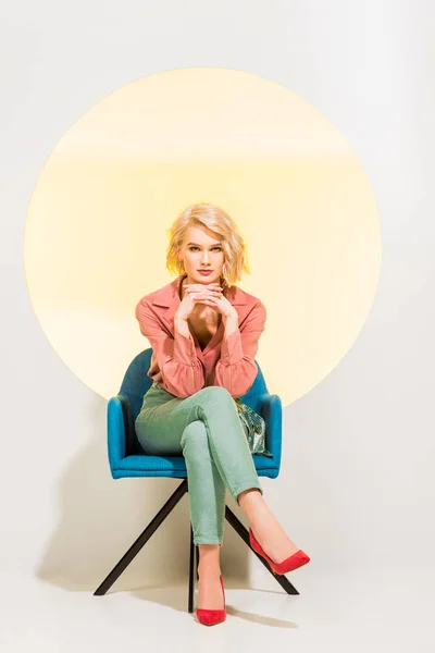 Belle fille élégante dans des vêtements colorés regardant l’appareil-photo et s’asseyant dans le fauteuil sur le blanc avec le cercle jaune — Photo de stock