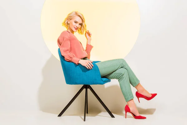 Belle fille élégante dans les vêtements colorés souriant tout en s’asseyant dans le fauteuil sur le blanc avec le cercle jaune — Photo de stock