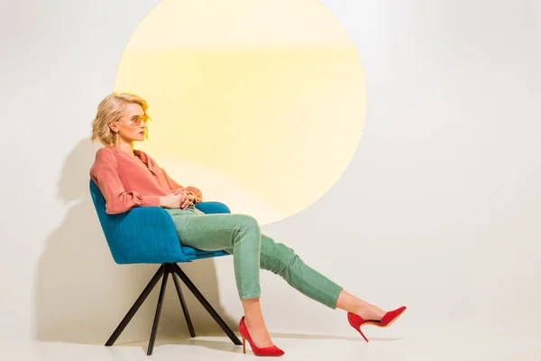 Hermosa chica de moda en ropa colorida sentado en sillón en blanco con círculo amarillo y espacio de copia - foto de stock